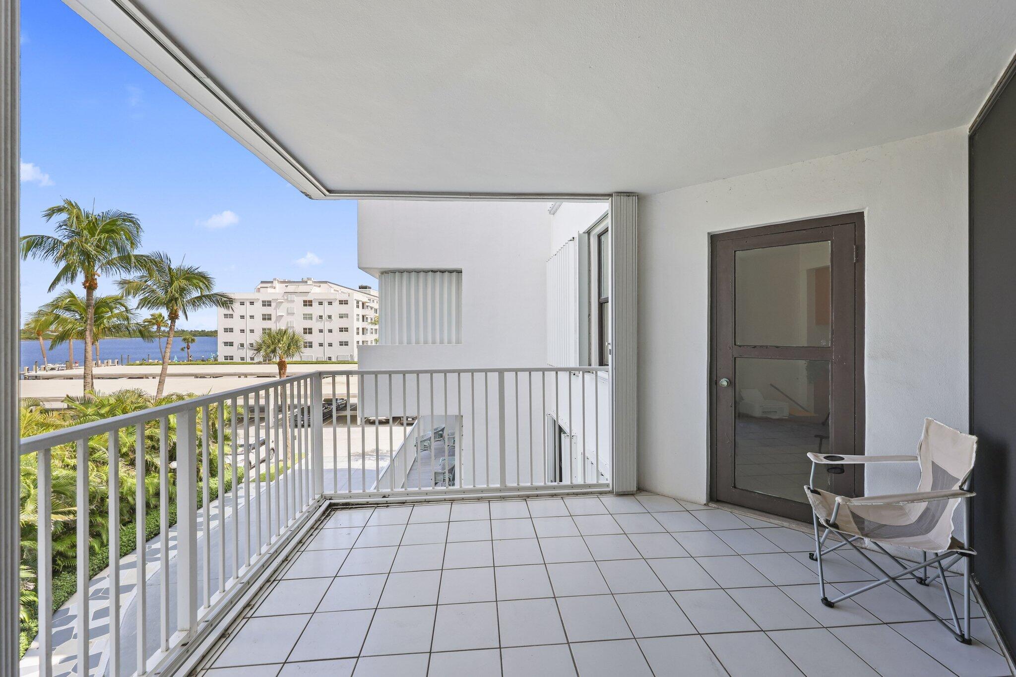 Apartment balcony with a tiled floor, folding chair, palm trees, and water view.