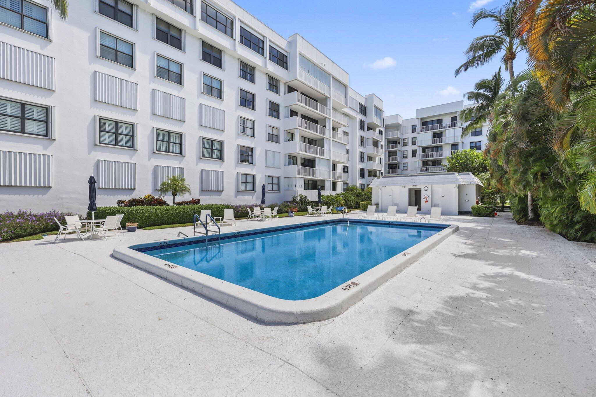 Condominium courtyard pool surrounded by white buildings, patio seating, and palm trees.