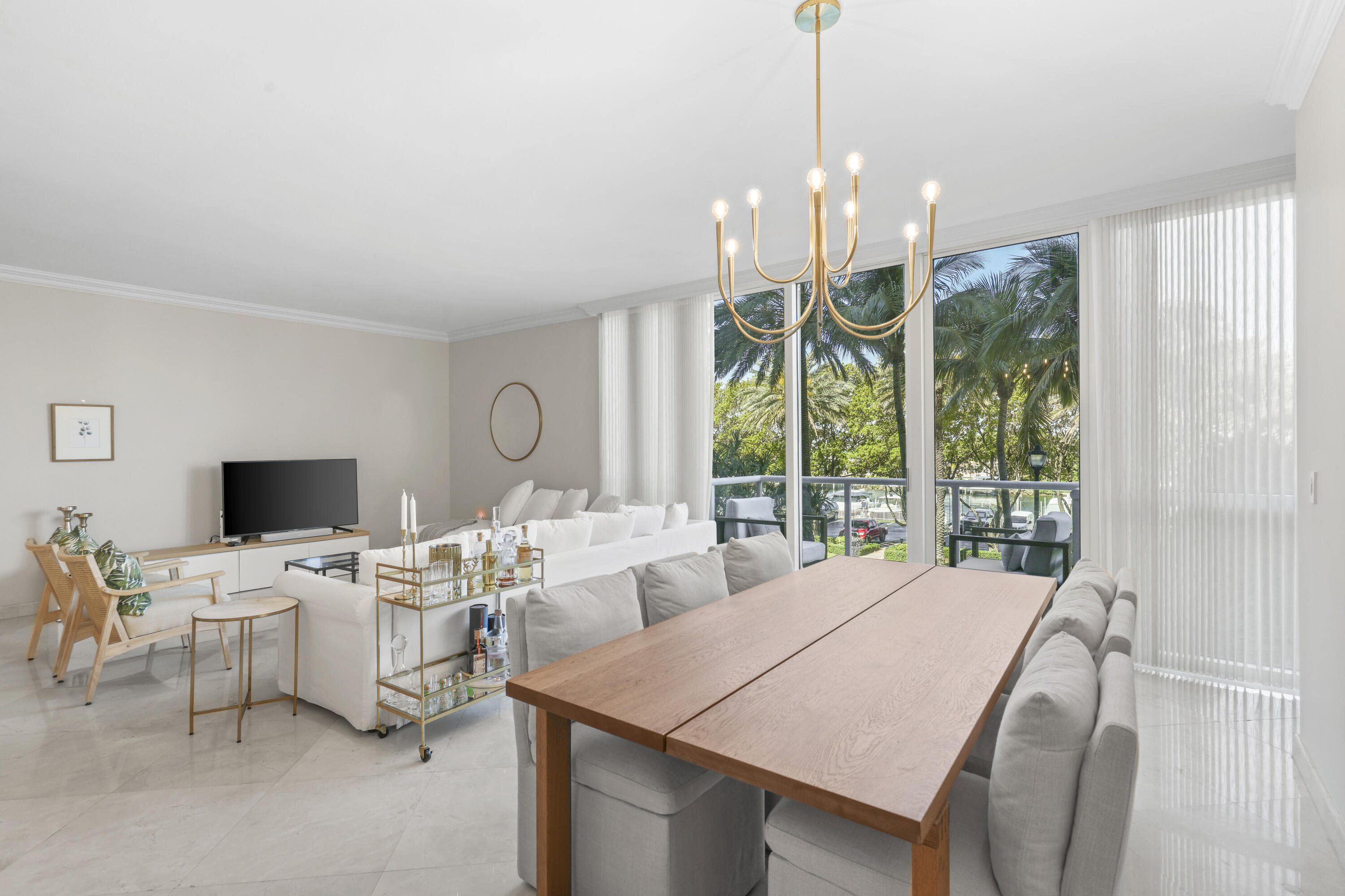 Open-plan living and dining room with a wooden table and palm trees visible through large windows.