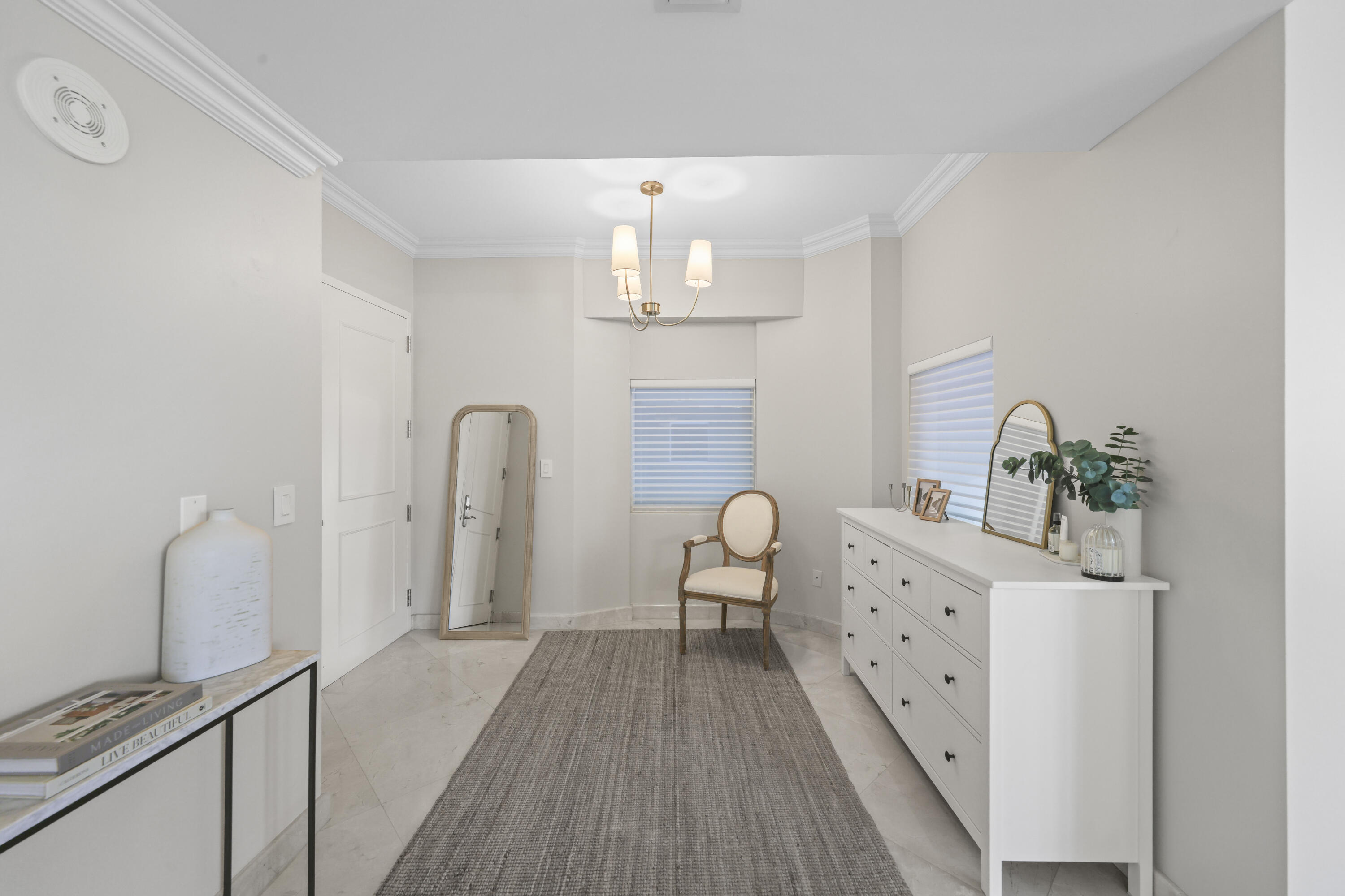 Dressing room with a long white dresser, upholstered chair, two mirrors, and windows with striped shades.
