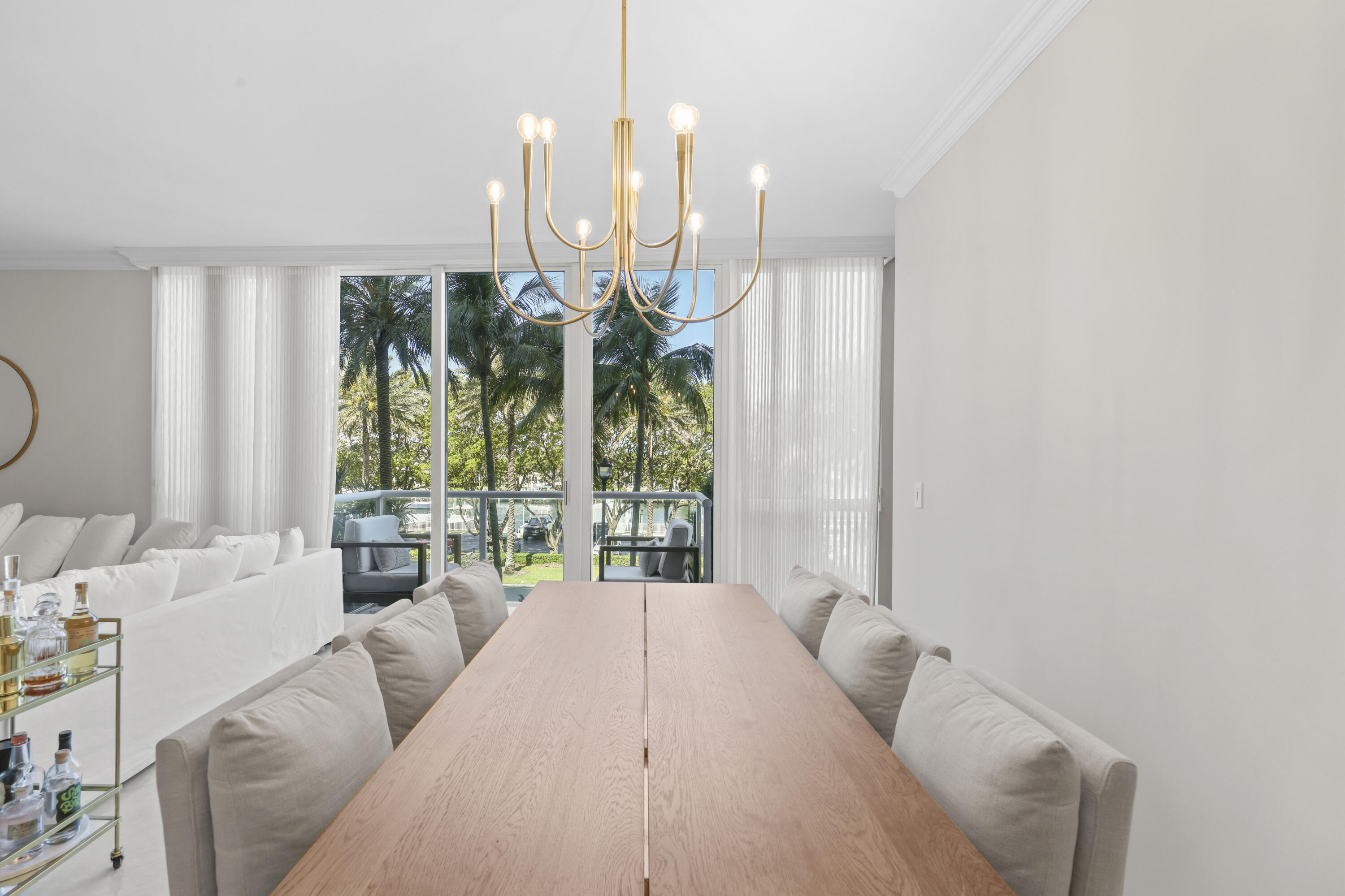 Dining area with a long wood table and gold chandelier, opening to a palm-lined balcony.