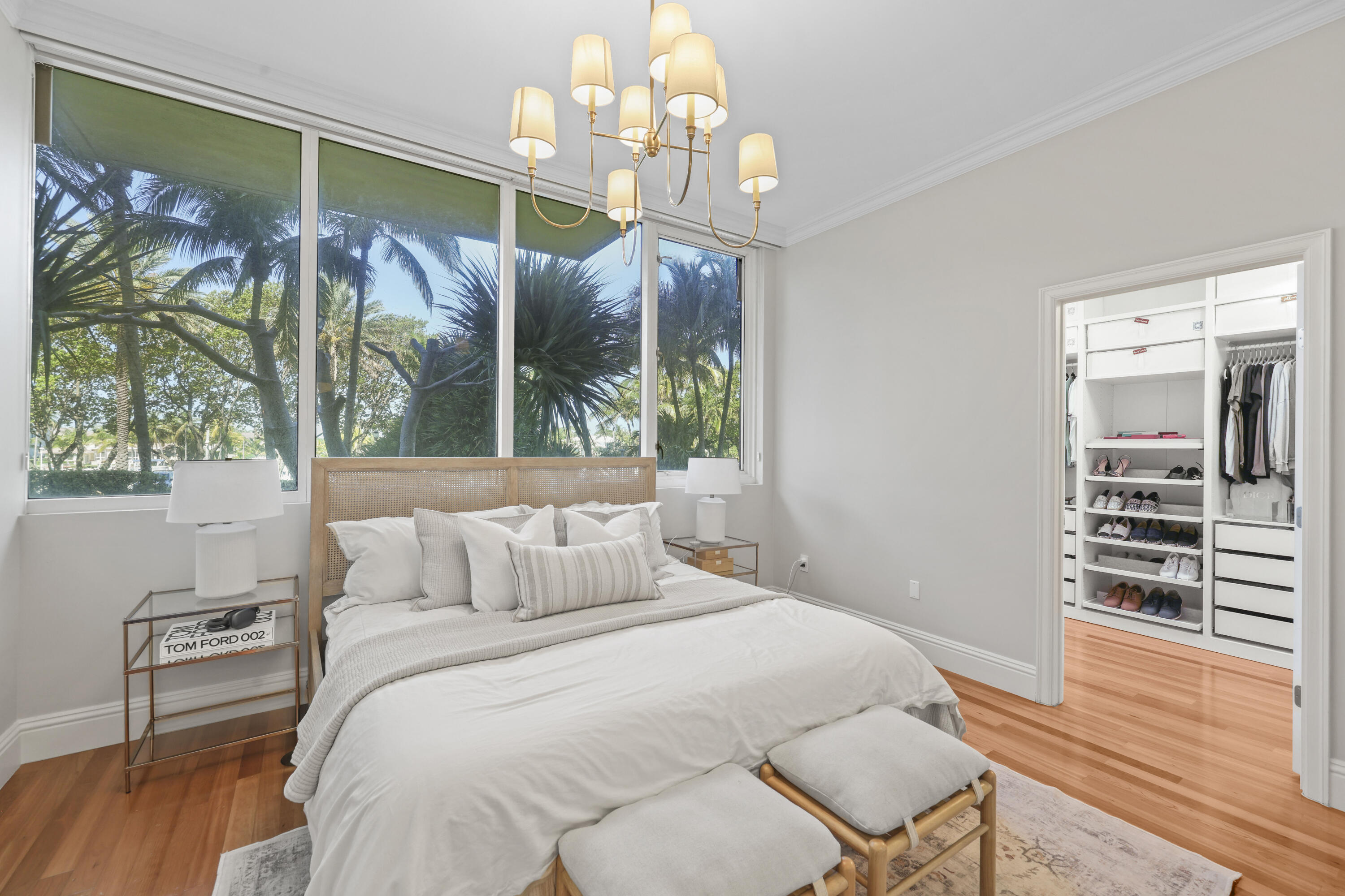 Bright bedroom with a white bed, palm trees outside large windows, and an open walk-in closet.