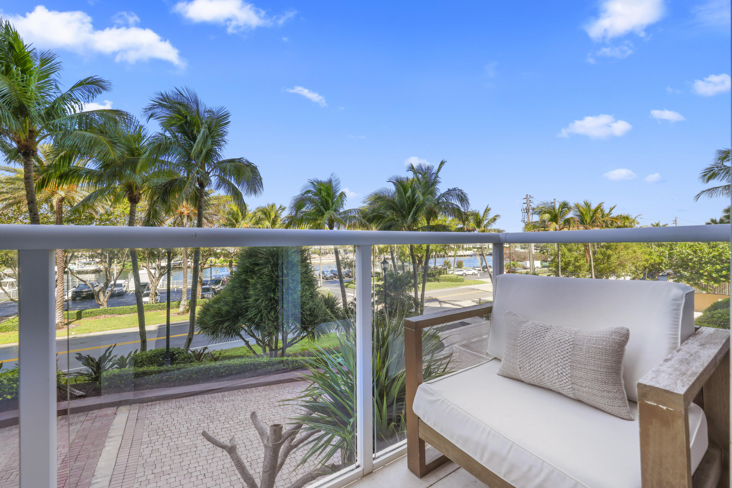 Furnished balcony overlooks a palm-lined canal and boats, with a white-cushioned wooden chair beside the glass railing.