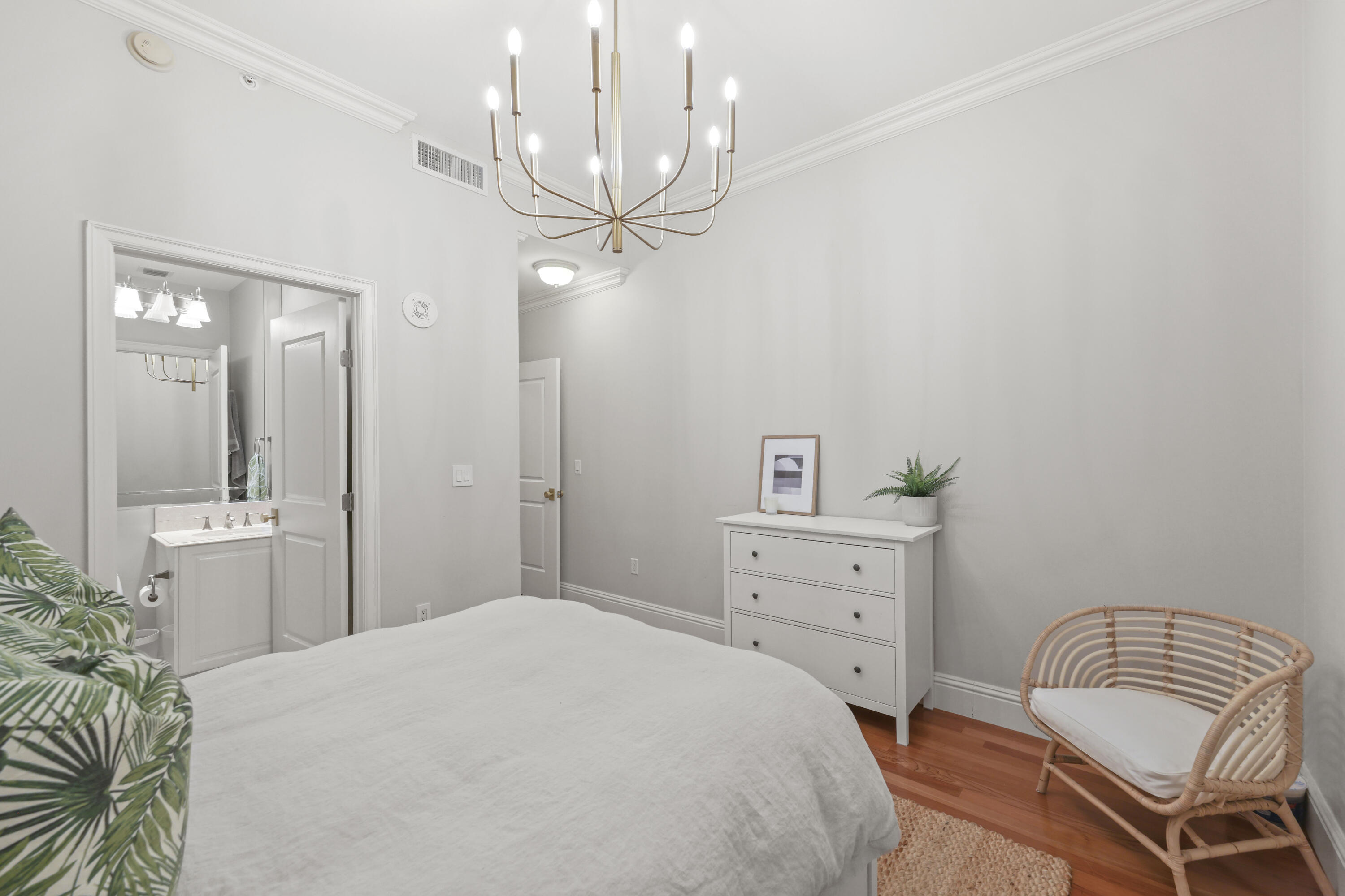 Bedroom with a brass chandelier, leafy pillows, white dresser, and adjoining bathroom vanity.