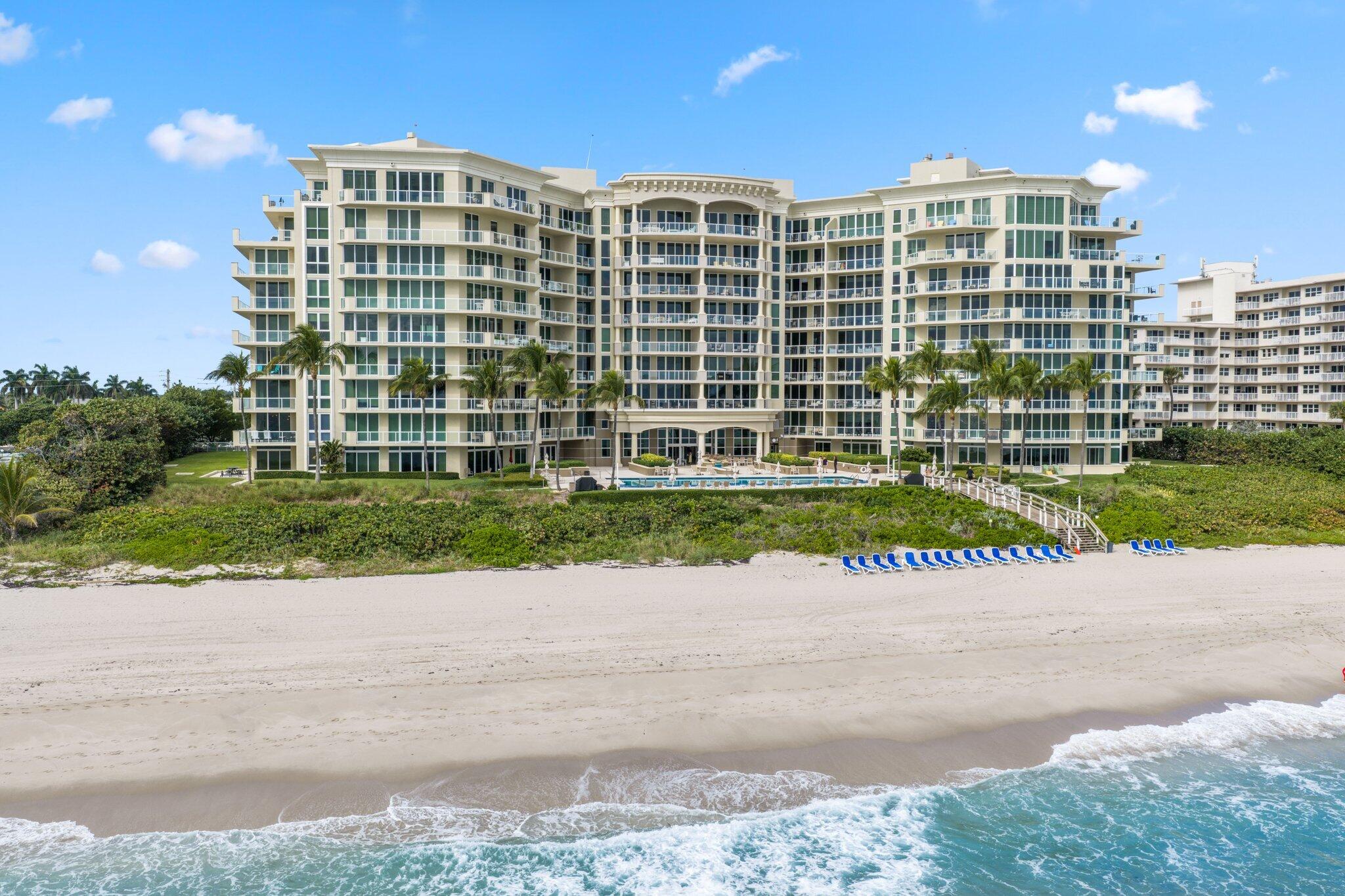 Beachfront condominium with glass balconies, a pool, palm trees, and blue lounge chairs along the shore.