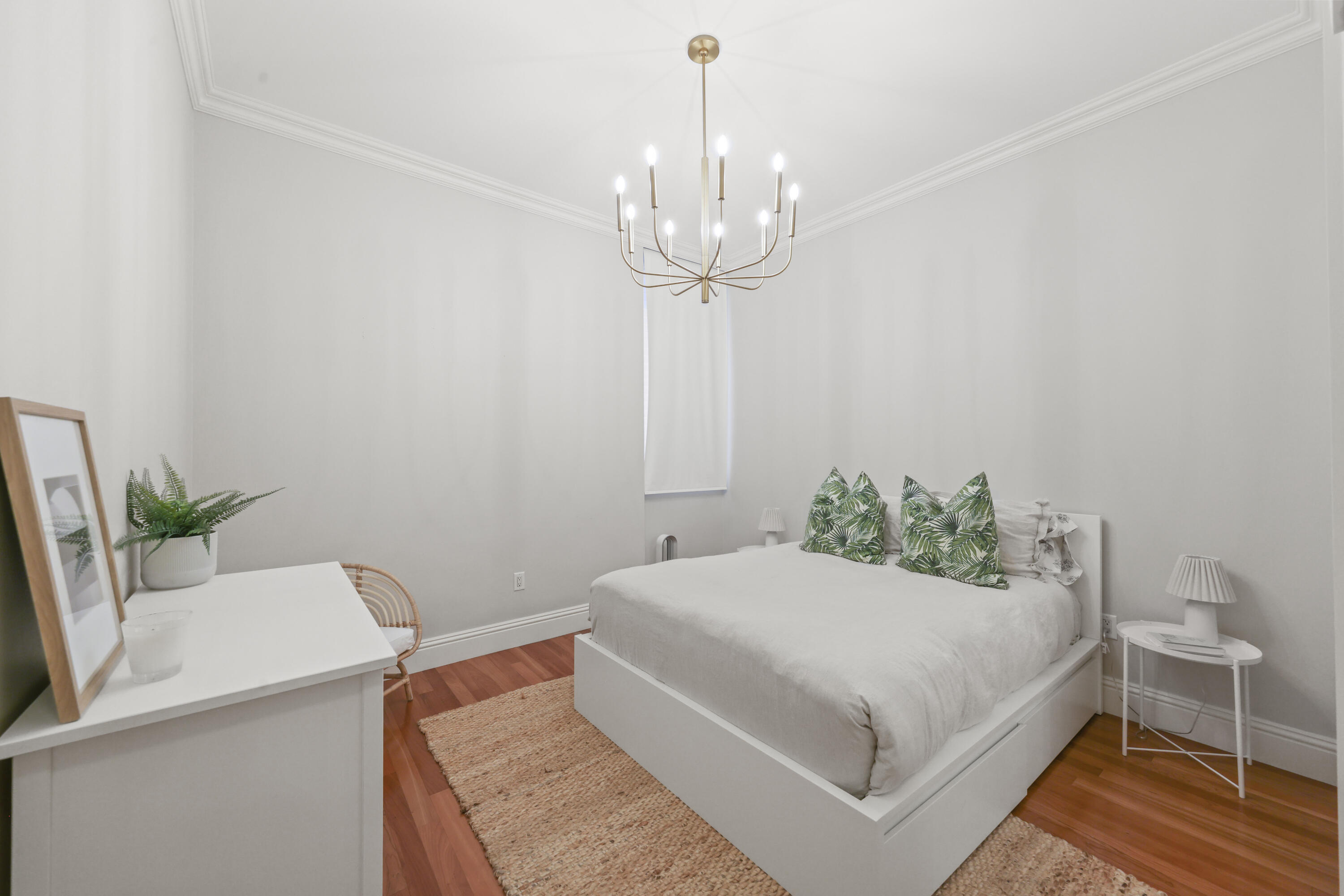 Bedroom with a white platform bed, tropical leaf-print pillows, and a brass chandelier.