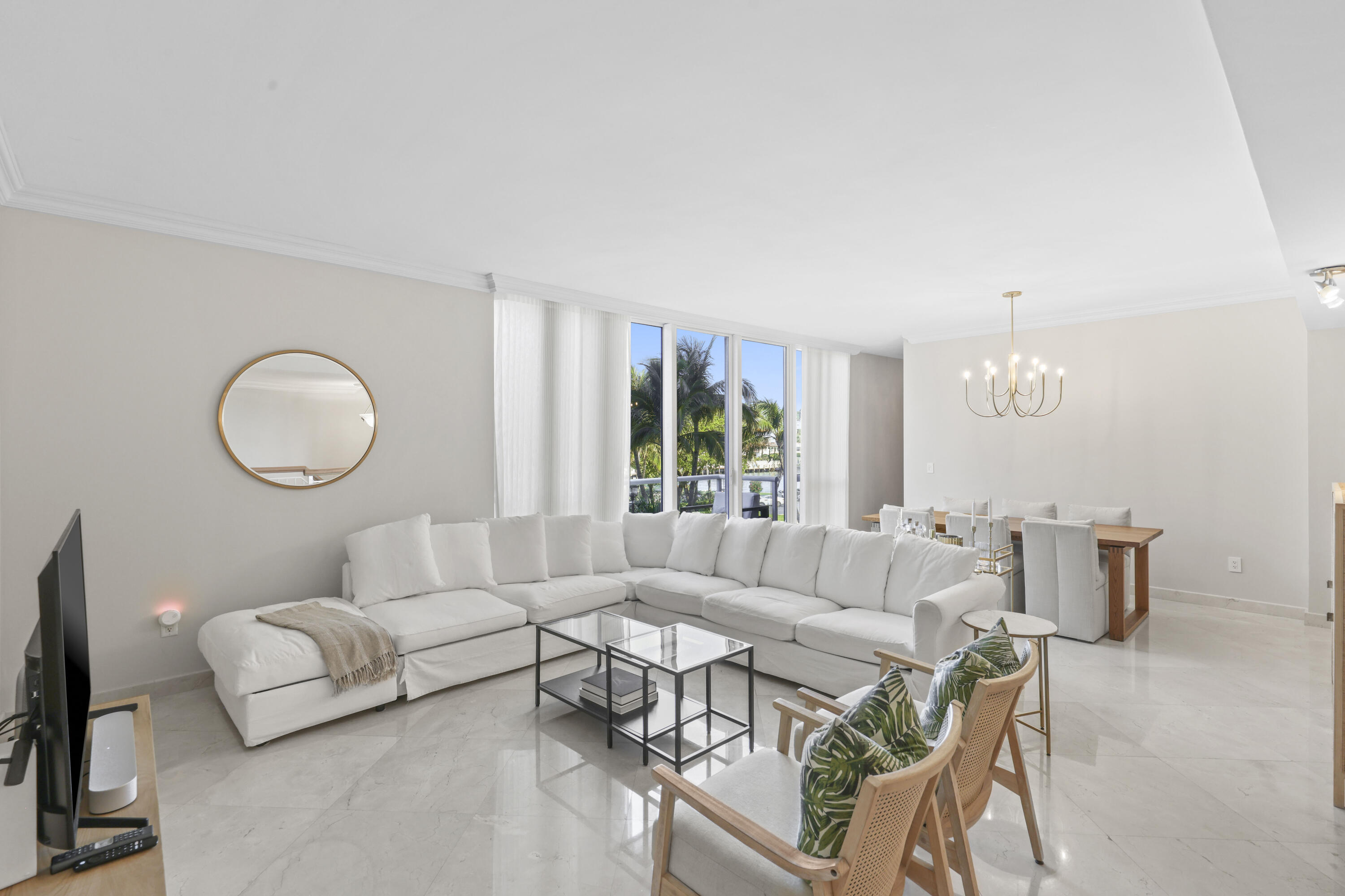 Living and dining room with a white sectional, glass coffee table, and windows overlooking palm trees.