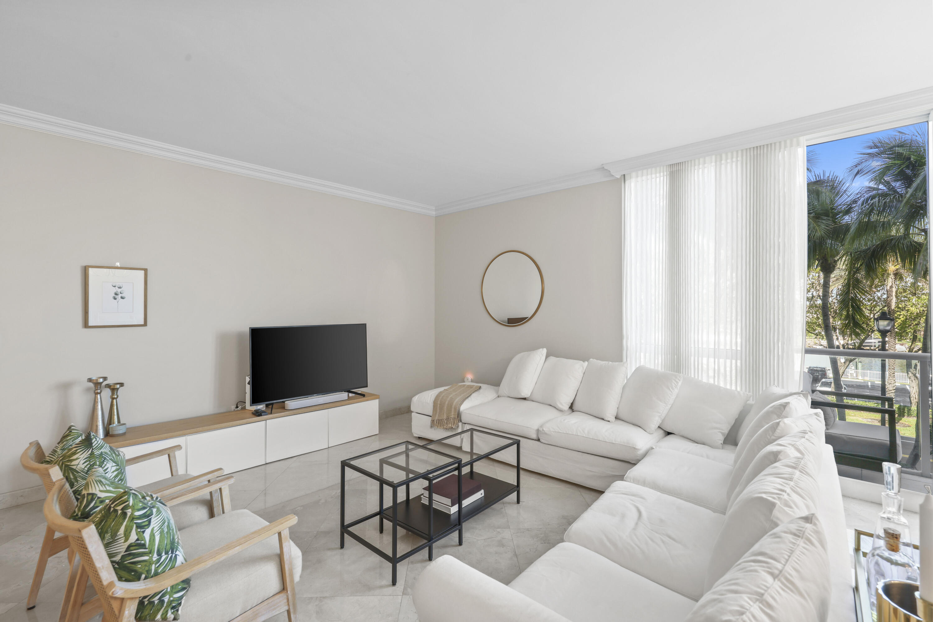 Furnished living room with white sectional sofas and palm trees visible through a large window.