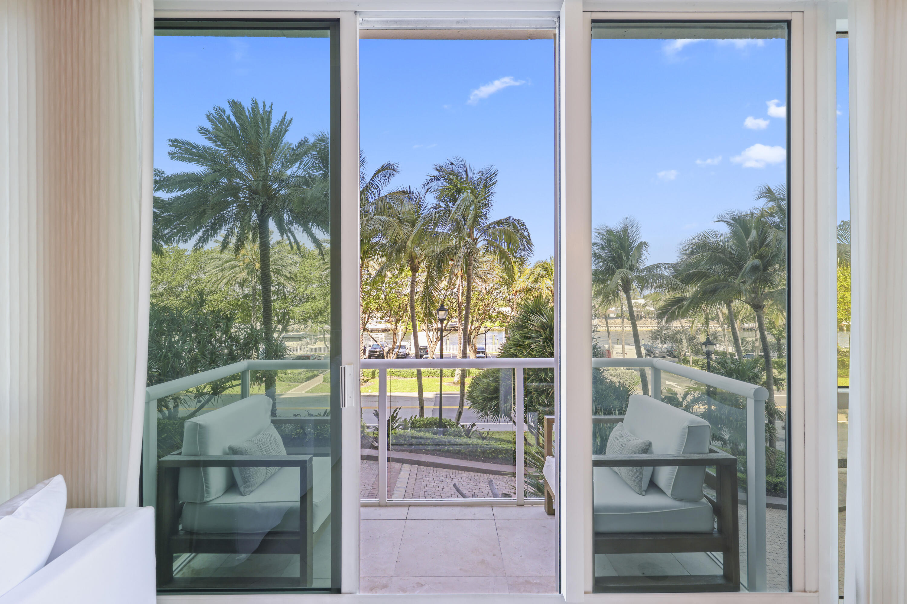 Condominium balcony with cushioned chairs overlooks palm trees and water through glass doors.
