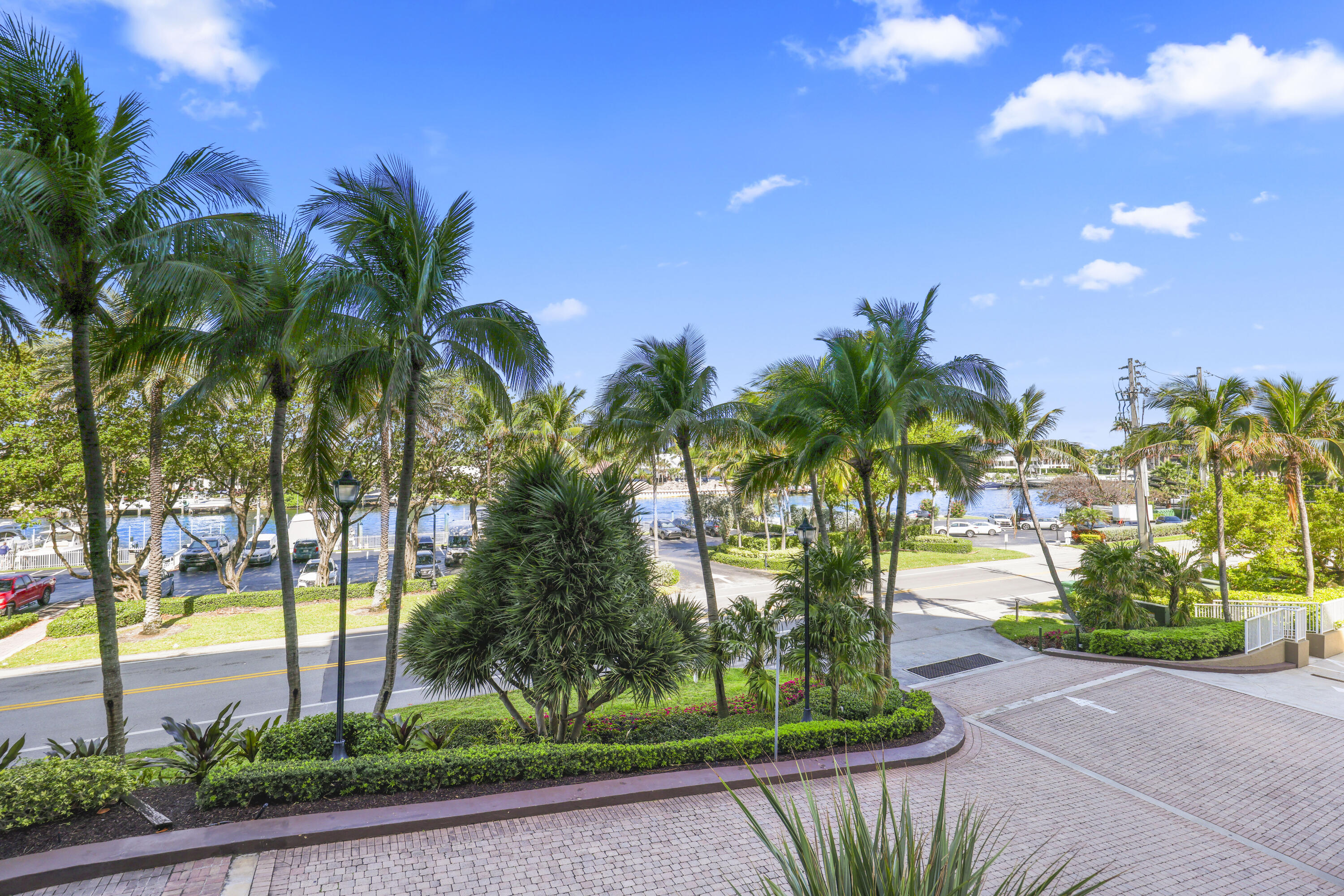 Palm-lined grounds and paved driveway overlook a road and marina canal with boats.