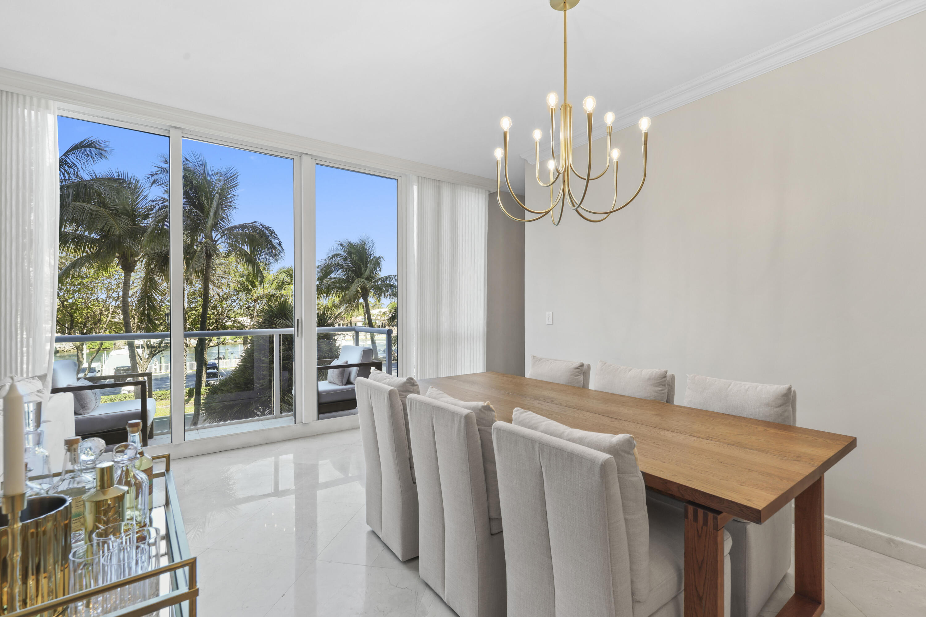 Dining room with a long wood table and gold chandelier, opening onto a palm-lined balcony with a water view.