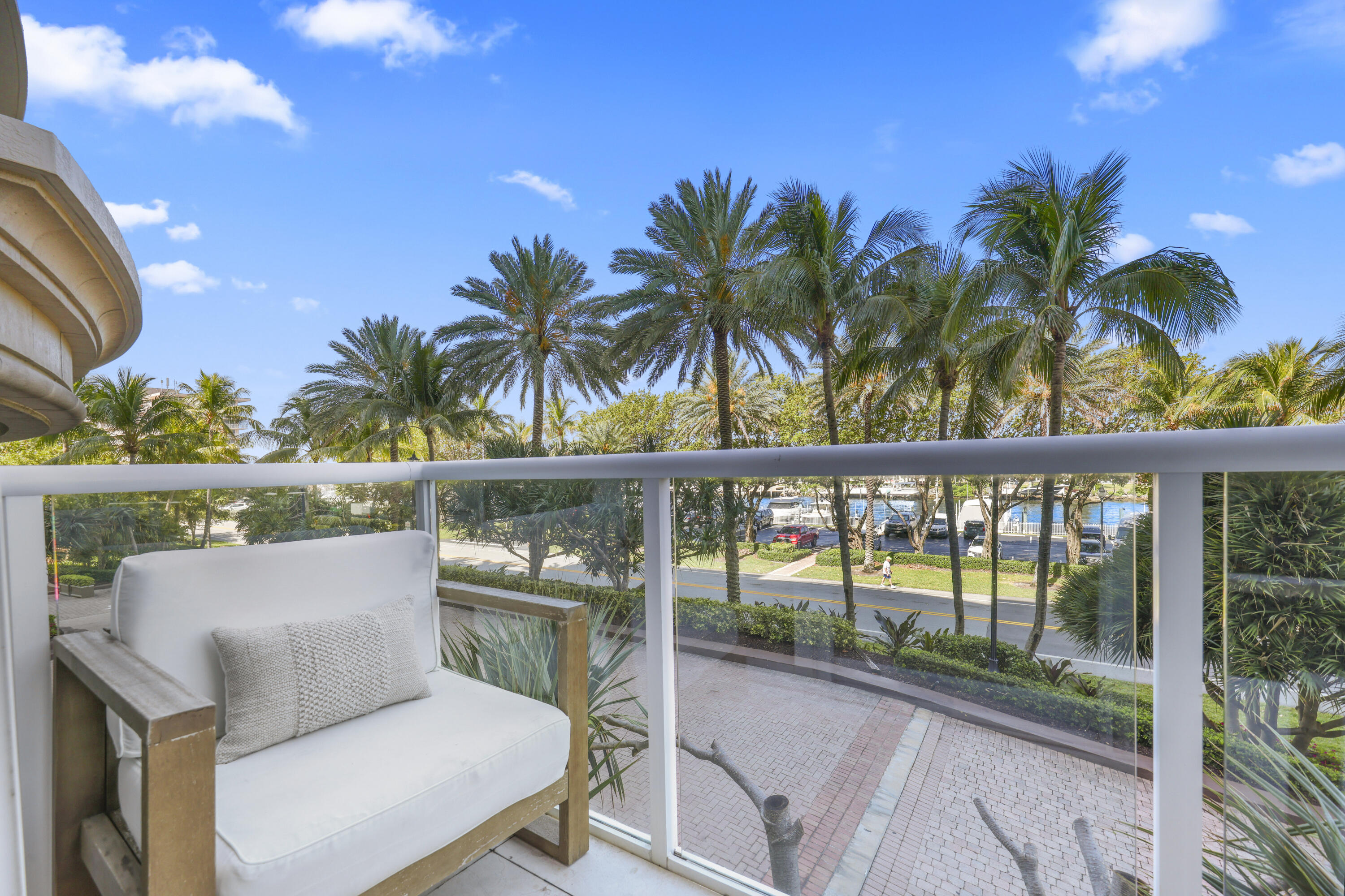 Furnished balcony overlooking palm trees, a street, and marina water, with a white-cushioned wooden chair.