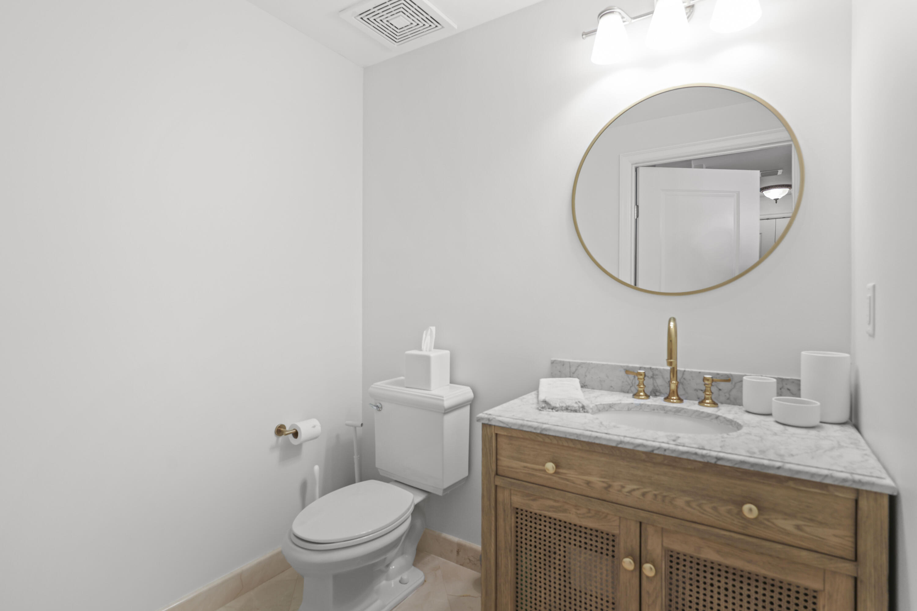 Bathroom with a wood vanity, marble countertop, gold fixtures, and a round mirror beside the toilet.