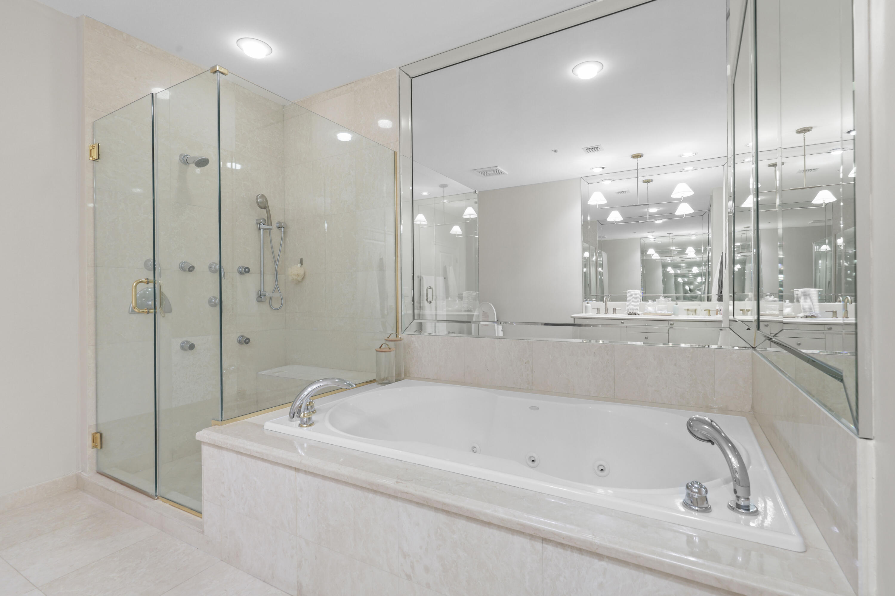 Bathroom with a large jetted tub, glass-enclosed shower, pale stone tile, and mirrored walls.
