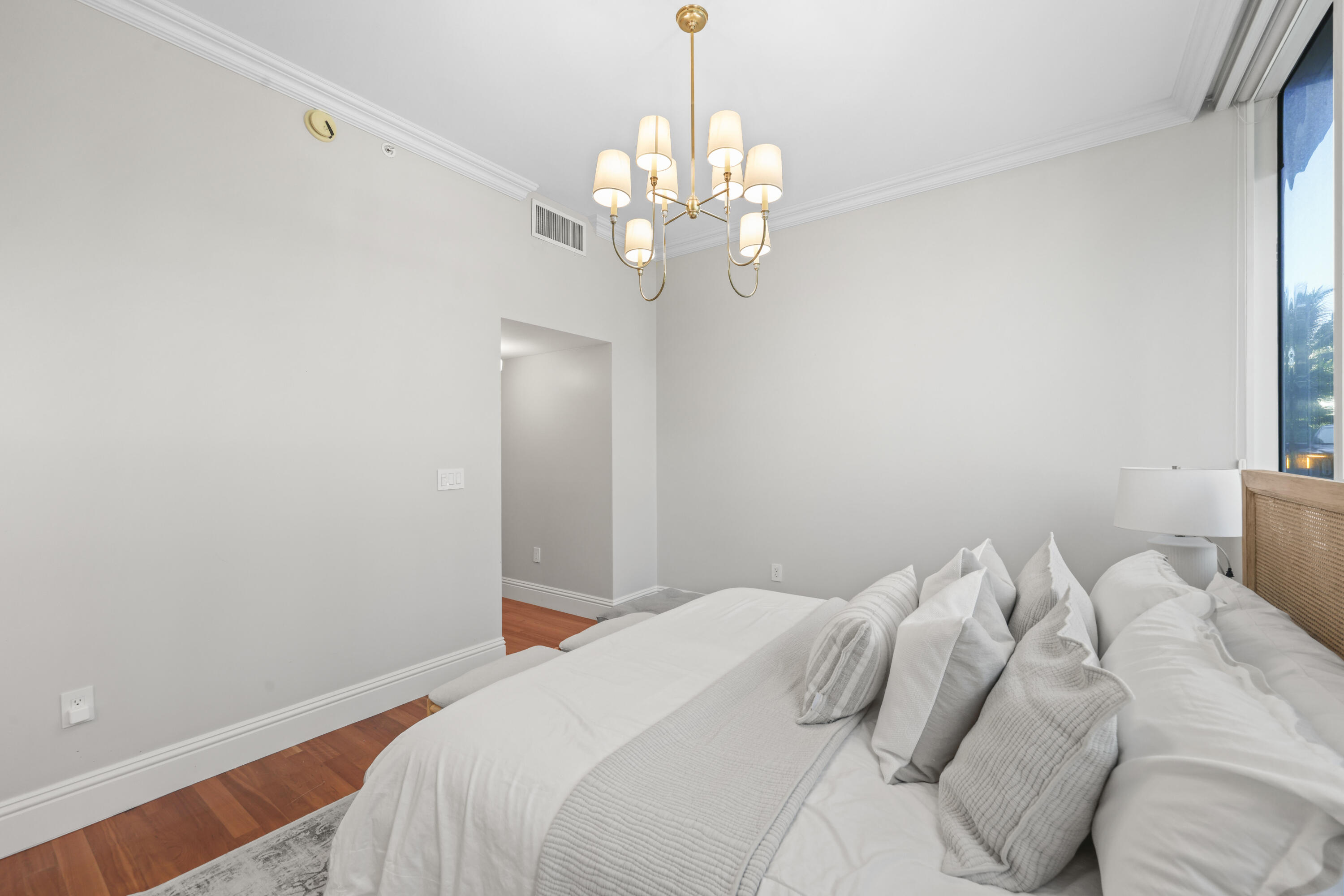 Bedroom with a white bed, layered pillows, brass chandelier, and warm wood floors.