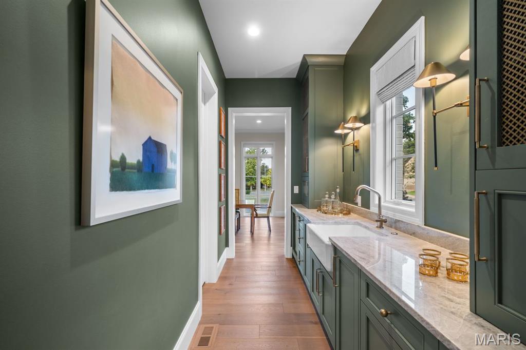 Narrow green kitchen with a farmhouse sink, marble-look counters, brass fixtures, and a bright dining area beyond.