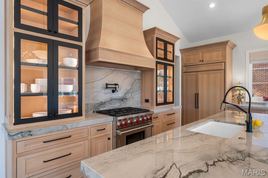Kitchen with light wood cabinetry, glass-front cabinets, a marble range hood, and a large marble island.