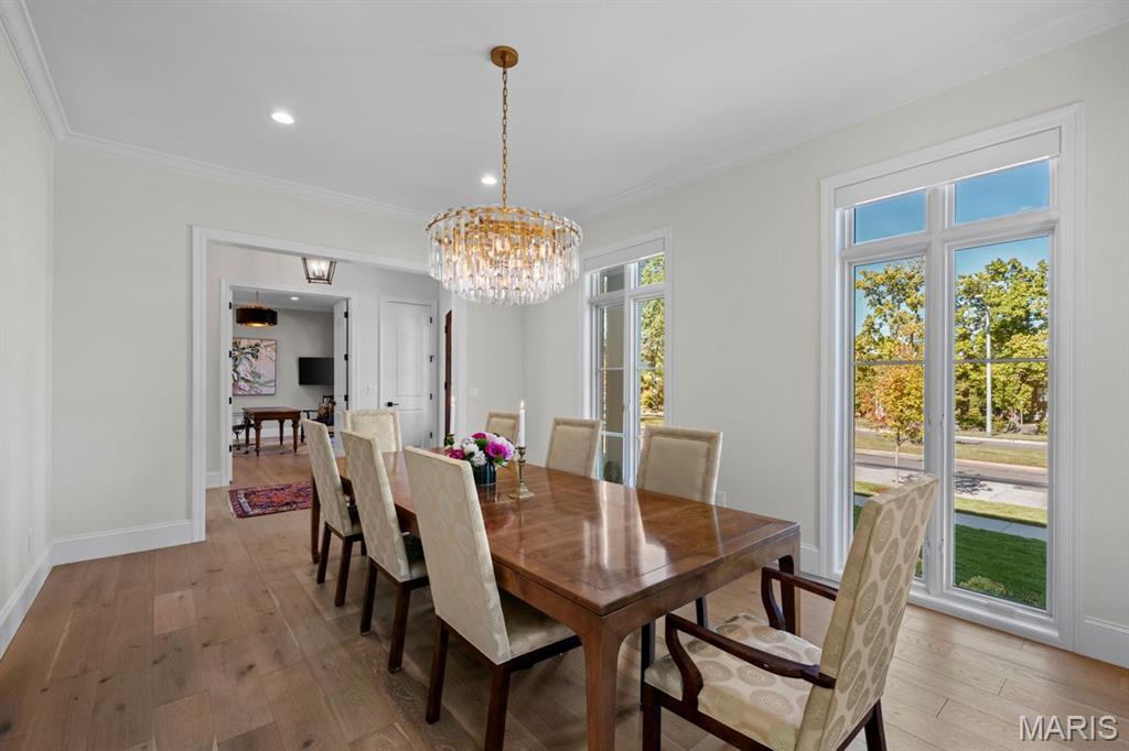 Dining room with a polished wood table, upholstered chairs, and a crystal chandelier beside tall windows.