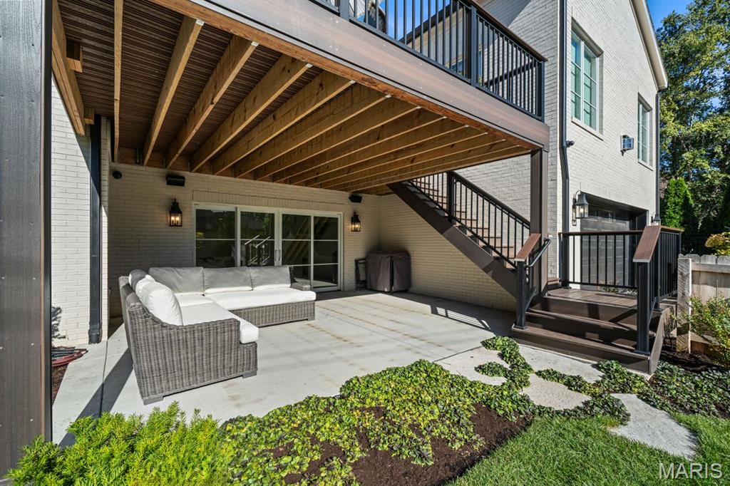Covered patio with wicker sectional seating, black metal stairs, and garden beds beside a white brick house.