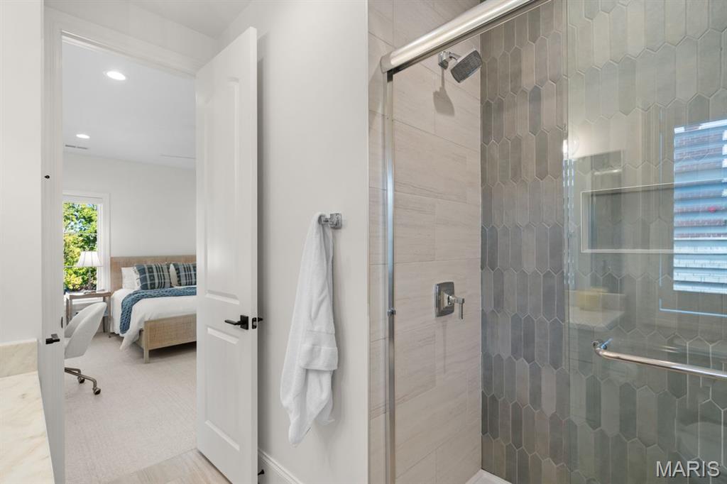 Bathroom with a glass shower featuring gray hexagonal tile, with a furnished bedroom visible through the doorway.