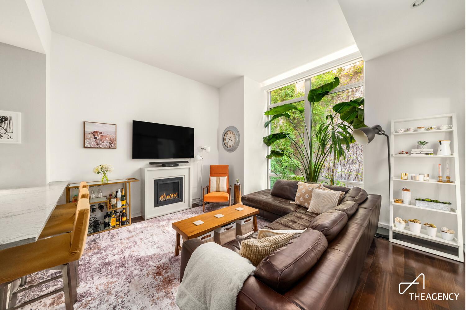 Living room with a brown leather sectional, fireplace, and large windows overlooking greenery.