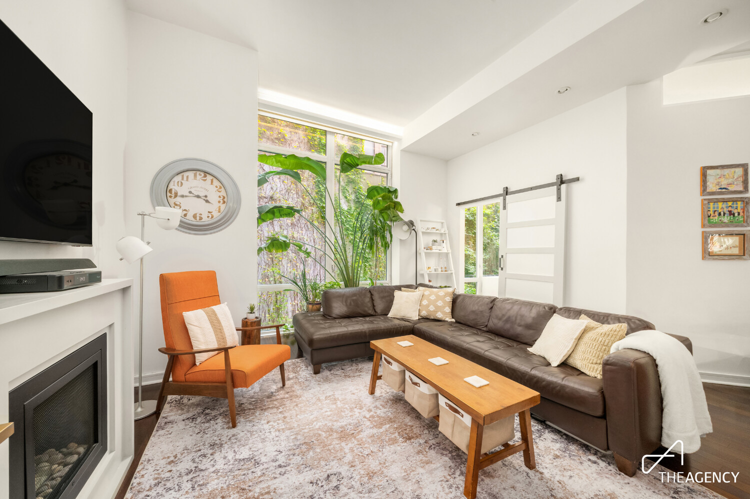 Living room with a brown leather sectional, orange chair, fireplace, and tall plant by large windows.