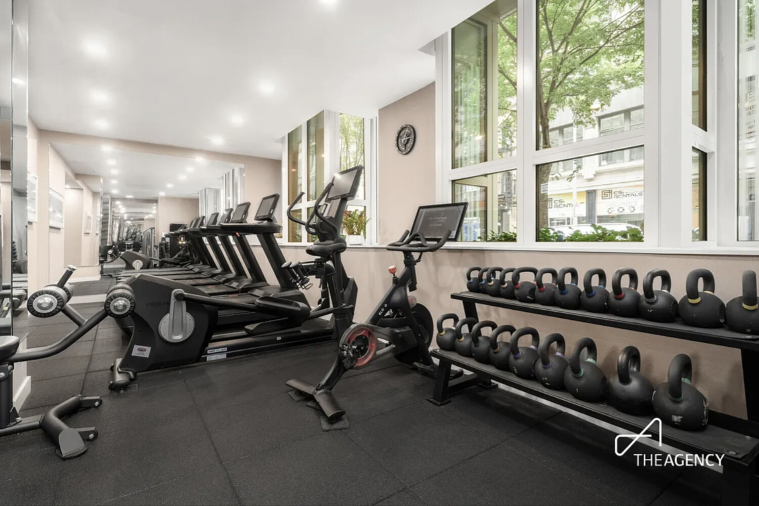 Building gym with treadmills and stationary bikes beside large windows and rows of kettlebells.