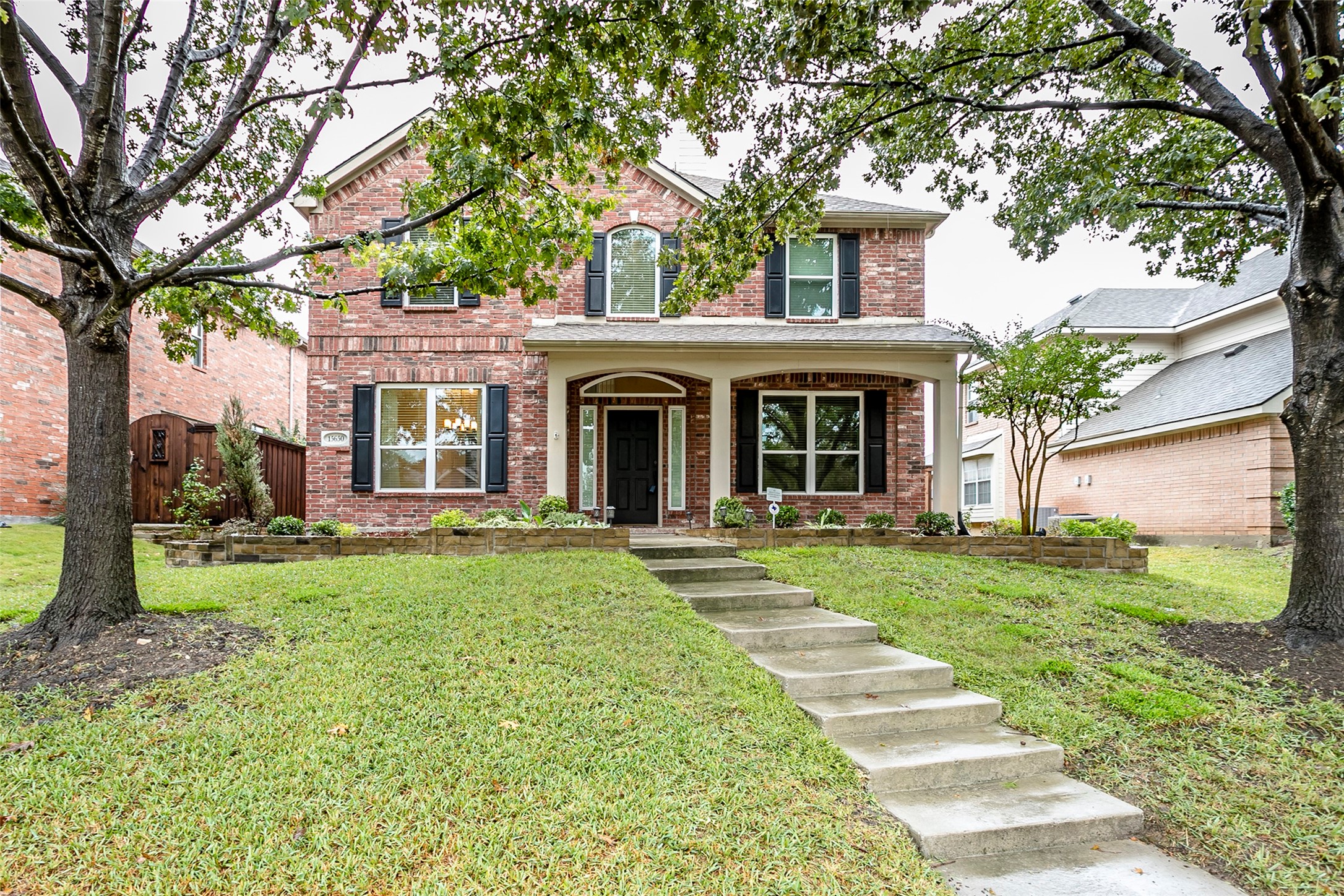 Two-story brick house with a covered porch, front walkway, lawn, and mature trees.