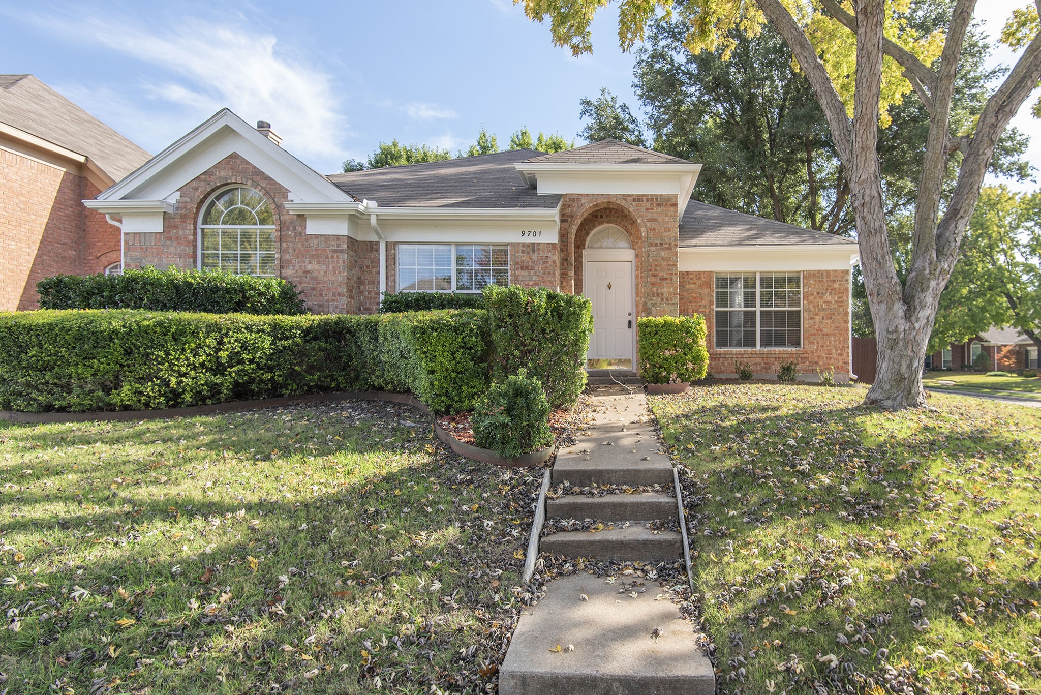 Red brick house with an arched front window, trimmed hedges, and a large tree beside the walkway.