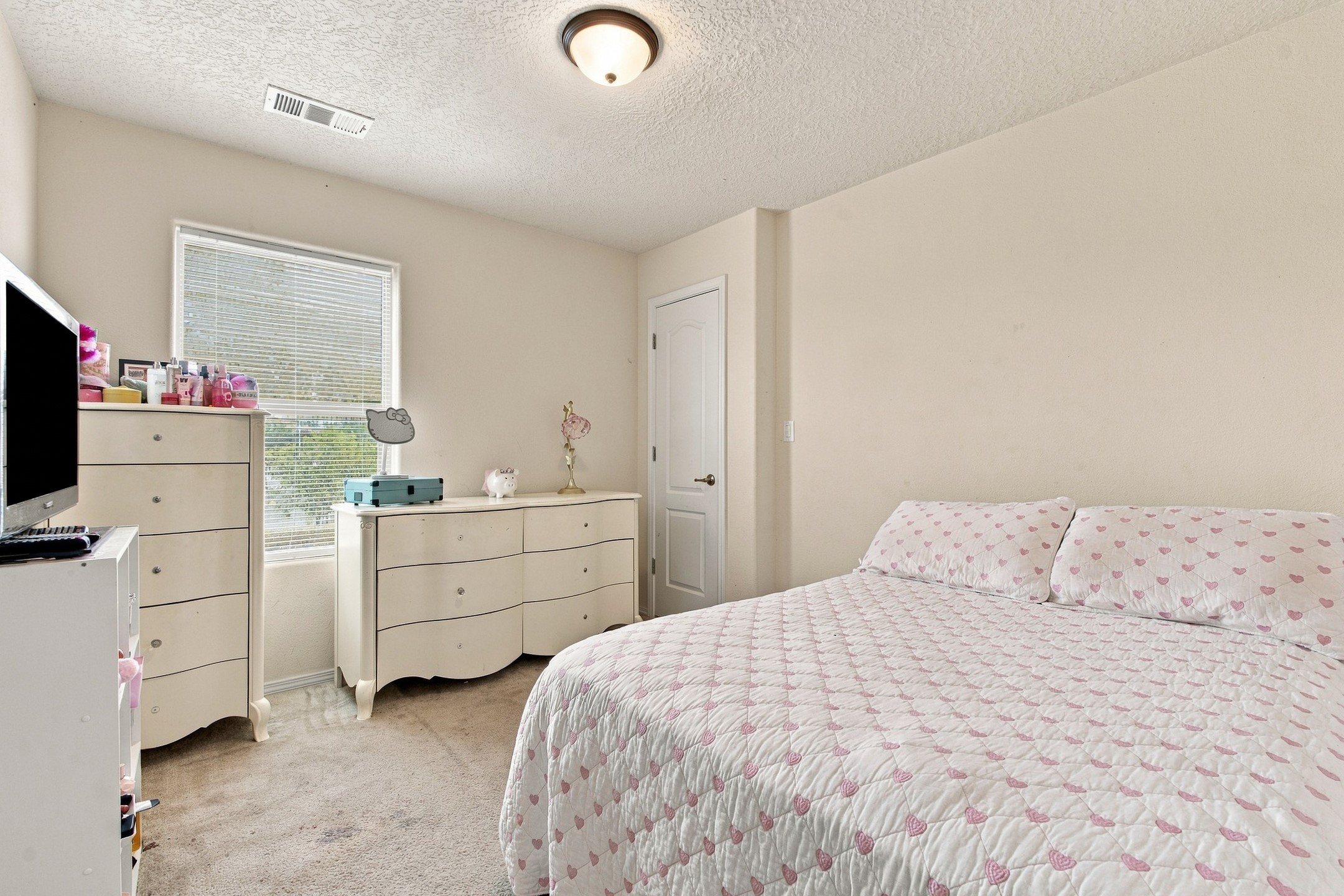 Bedroom with a pink heart-patterned quilt, cream dressers, and a window with blinds.