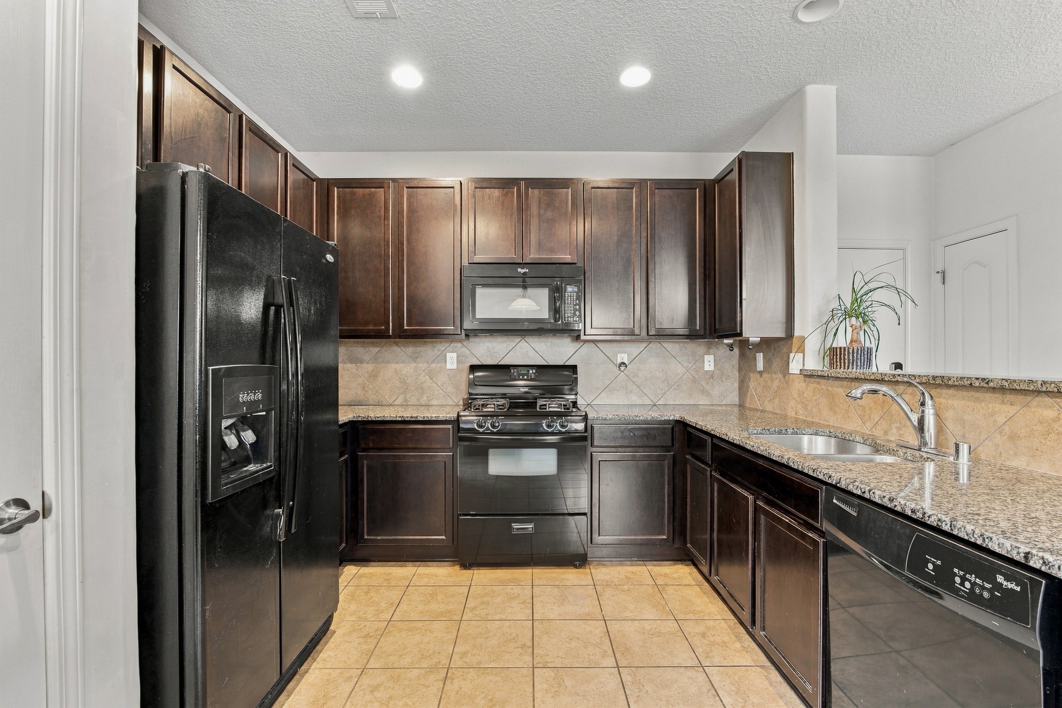 Kitchen with dark wood cabinets, granite counters, black appliances, and tan tile flooring.