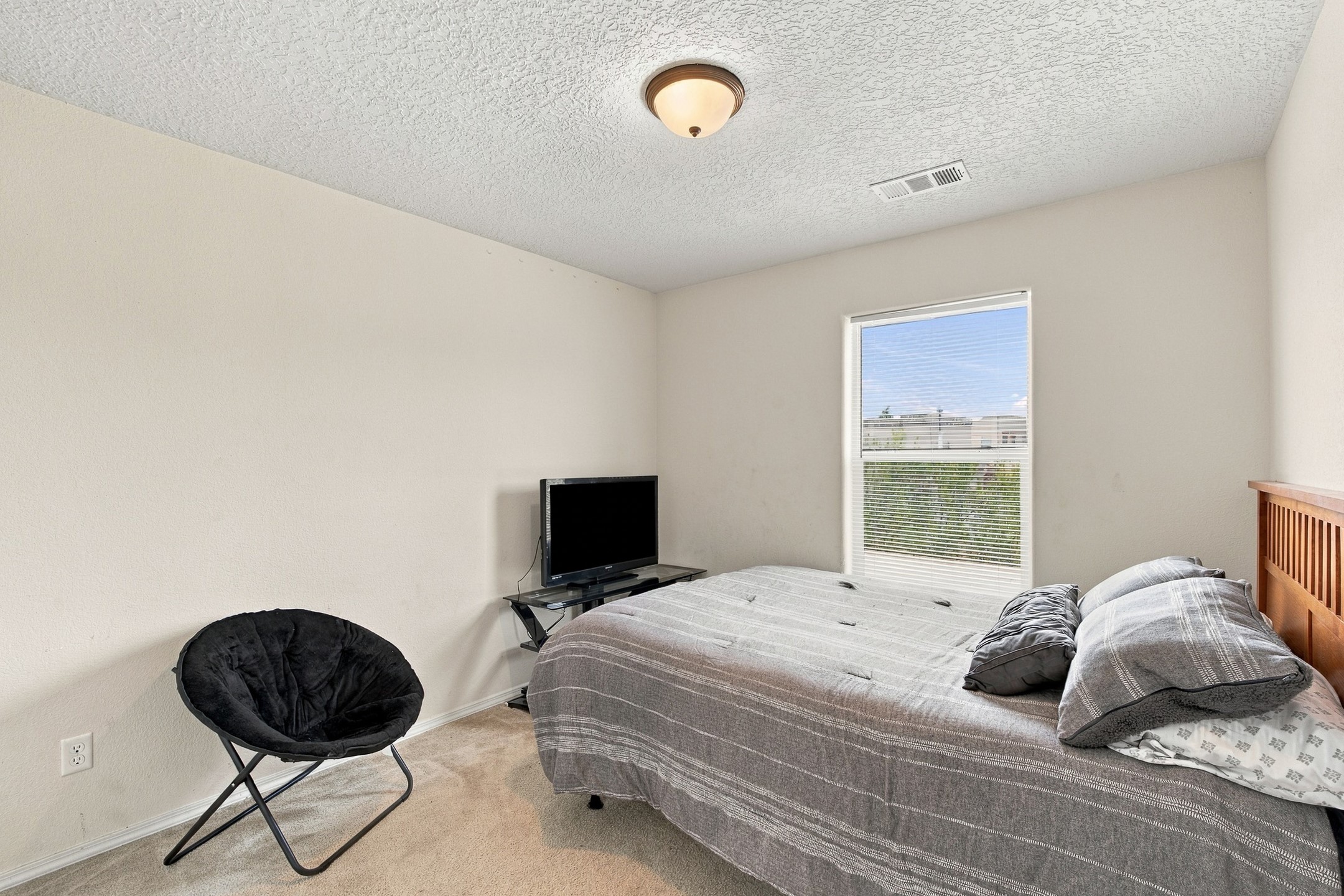 Bedroom with a gray-covered bed, black lounge chair, and television beside a window.