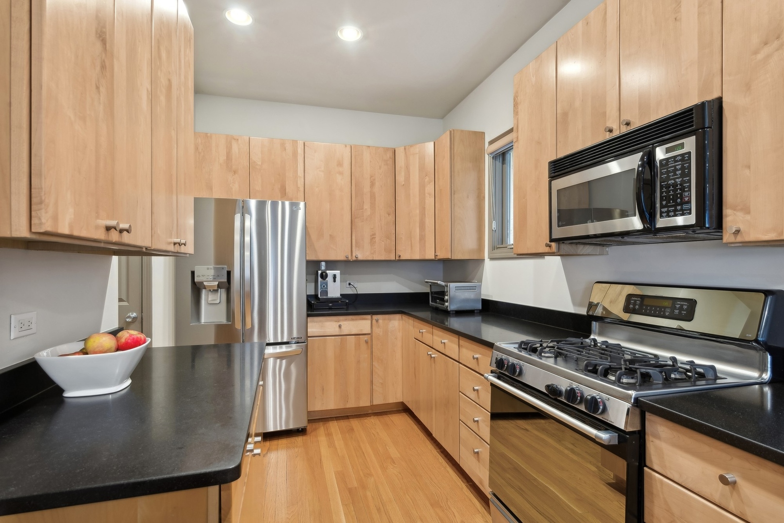 Kitchen with light wood cabinets, black countertops, stainless steel appliances, and hardwood floors.