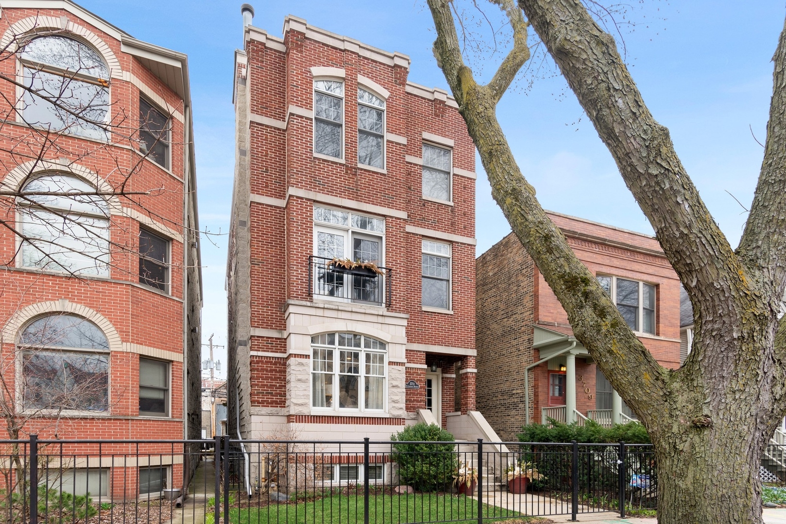 Red brick multi-story residence with arched windows, a small balcony, and a fenced front garden.