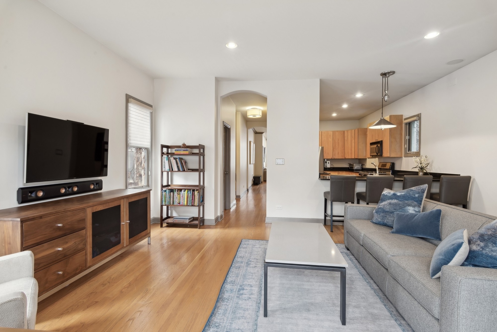 Open-plan living room and kitchen with hardwood floors, gray sofa, blue accents, and warm wood cabinetry.