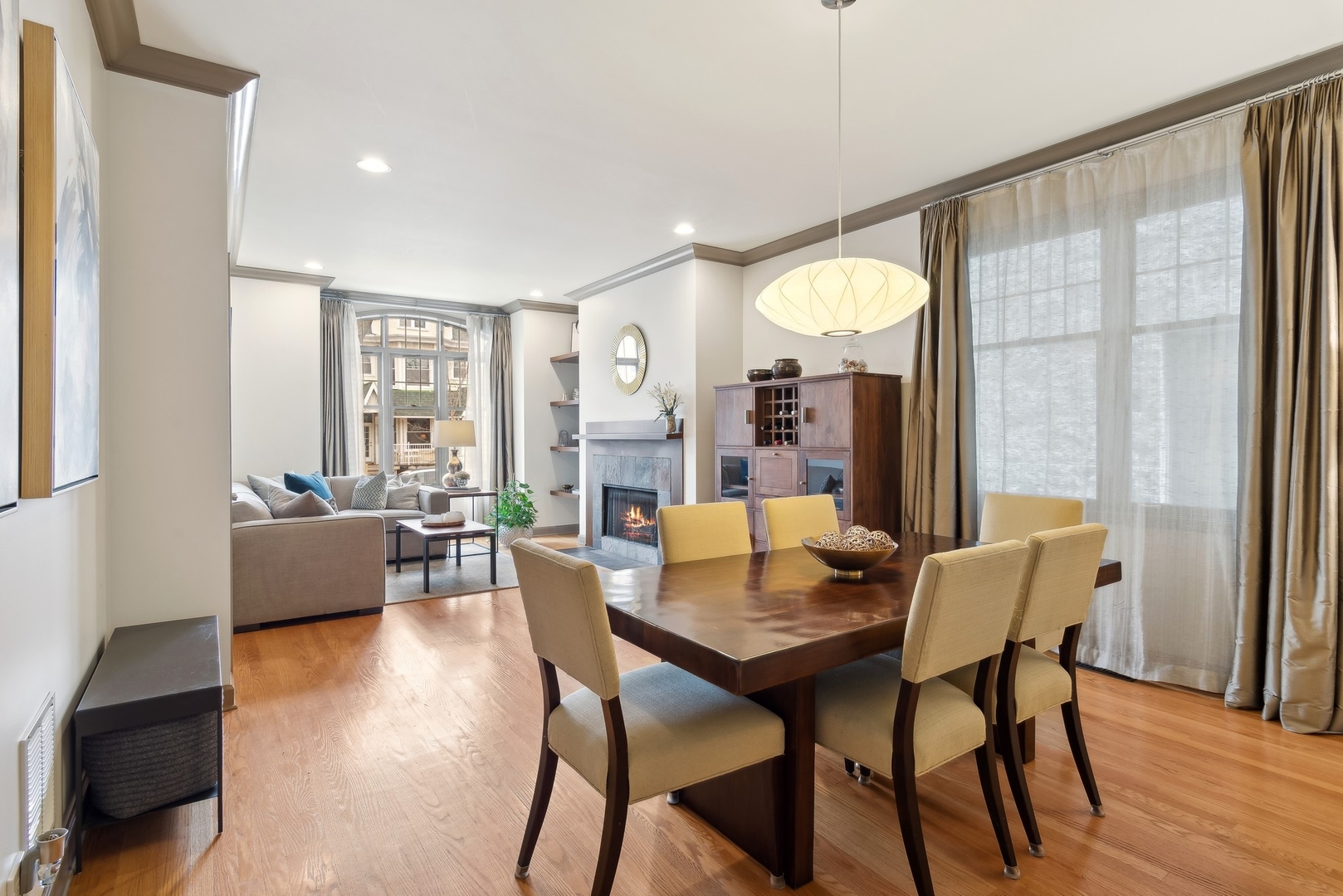 Dining and living room with a wood table, fireplace, hardwood floors, and large curtained windows.