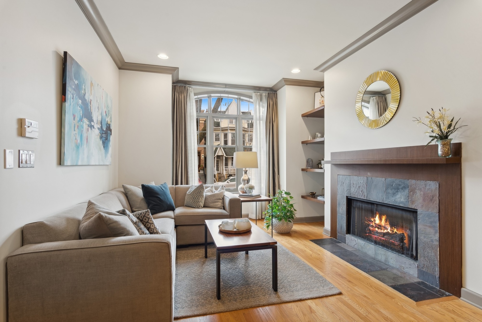 Staged living room with a sectional sofa and lit stone fireplace beside a large arched window.
