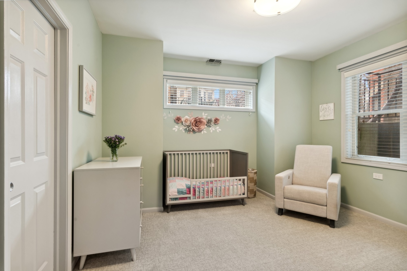 Furnished nursery with a gray crib, sage-green walls, floral decor, and a cream armchair.