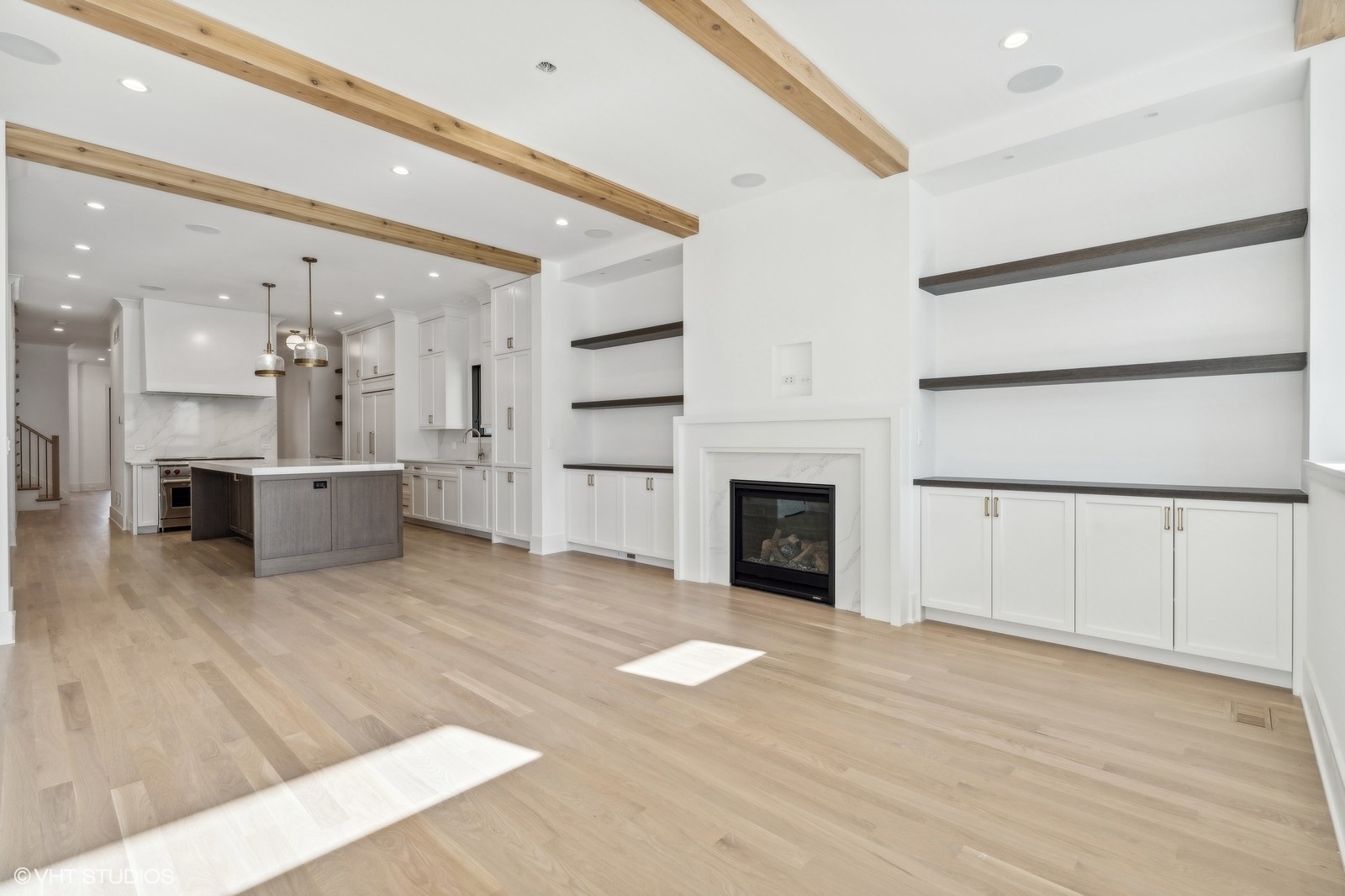 Open-plan living room and kitchen with exposed wood beams, a marble fireplace, and gray island.