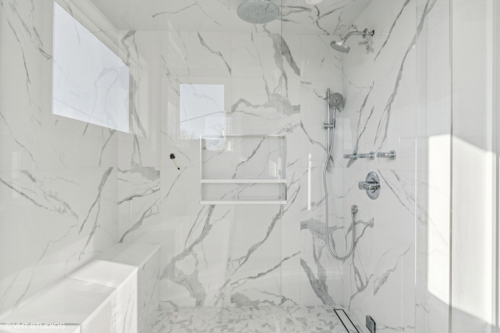 Walk-in shower with marble-look tile, built-in bench, recessed niche, and multiple showerheads.