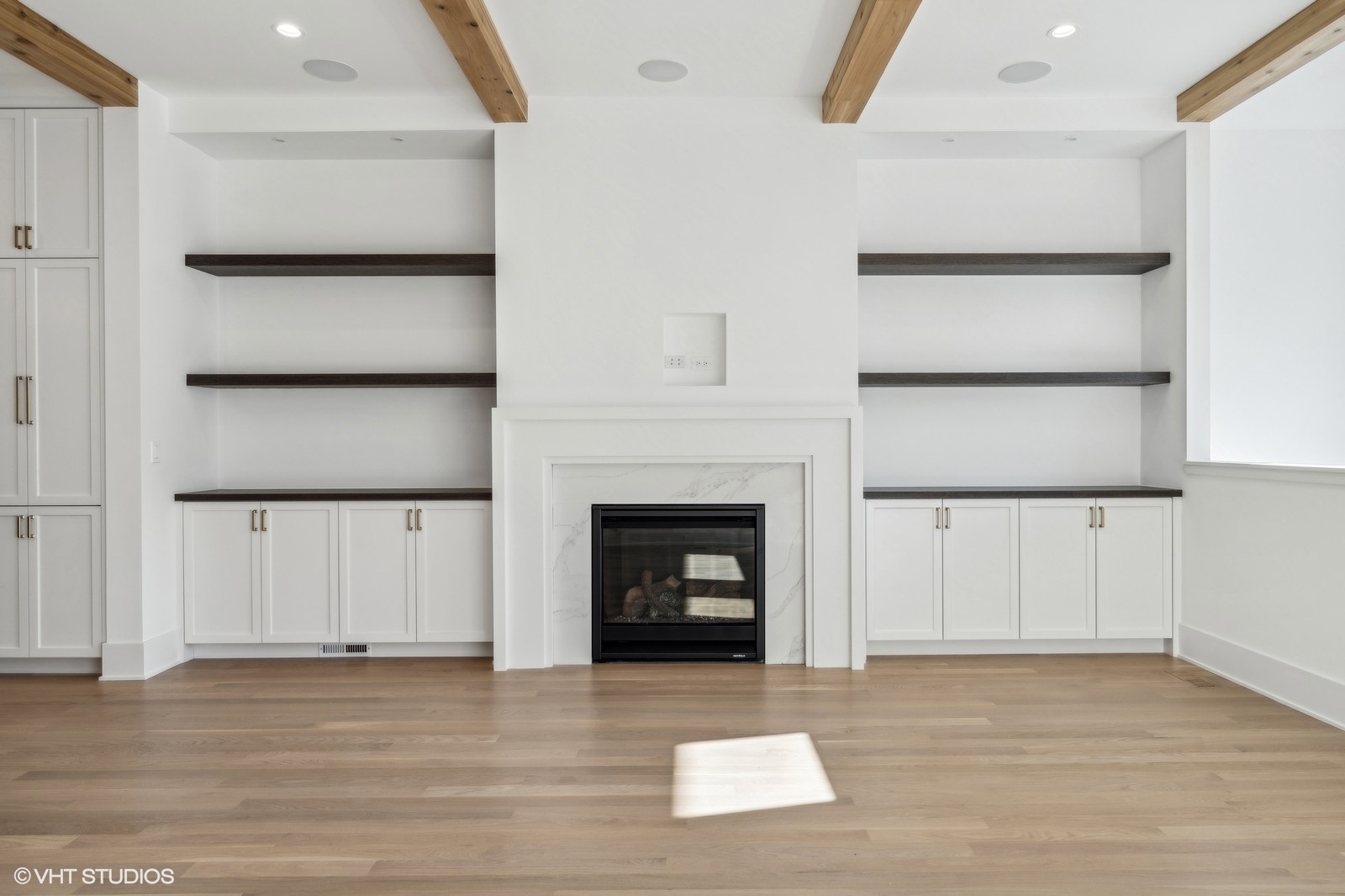 Vacant living room with a white fireplace, built-in shelves, exposed wood beams, and hardwood floors.