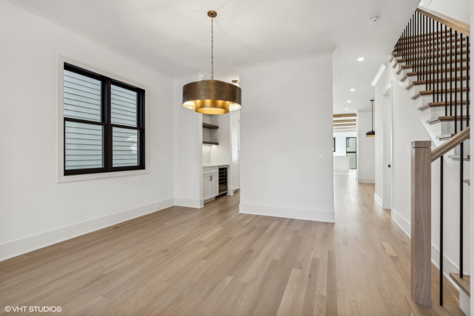 Empty dining area with light wood floors, bronze pendant chandelier, and staircase with dark balusters.