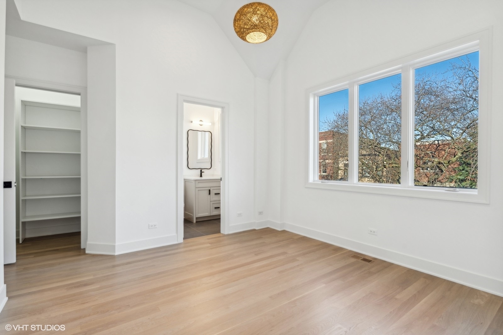 Vacant bedroom with vaulted ceiling, large windows, light hardwood floors, and an adjoining bathroom and closet.