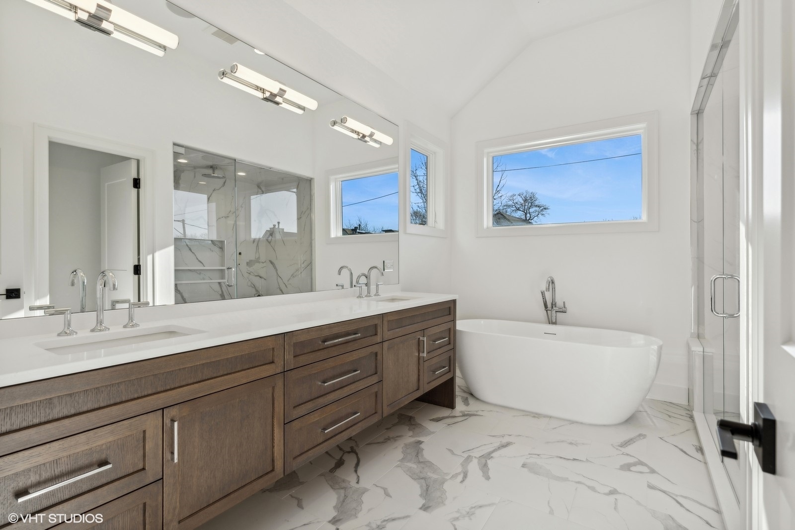 Bright bathroom with a dark wood double vanity, freestanding tub, and marble-patterned floors.