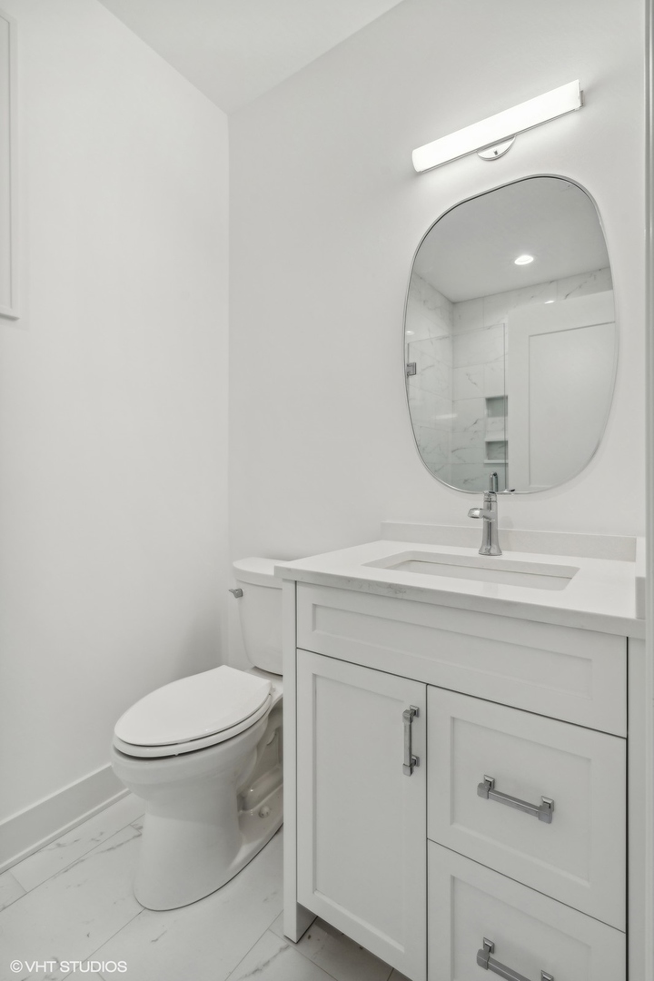 White bathroom vanity and toilet with an oval mirror and marble-look shower tile reflected behind them.