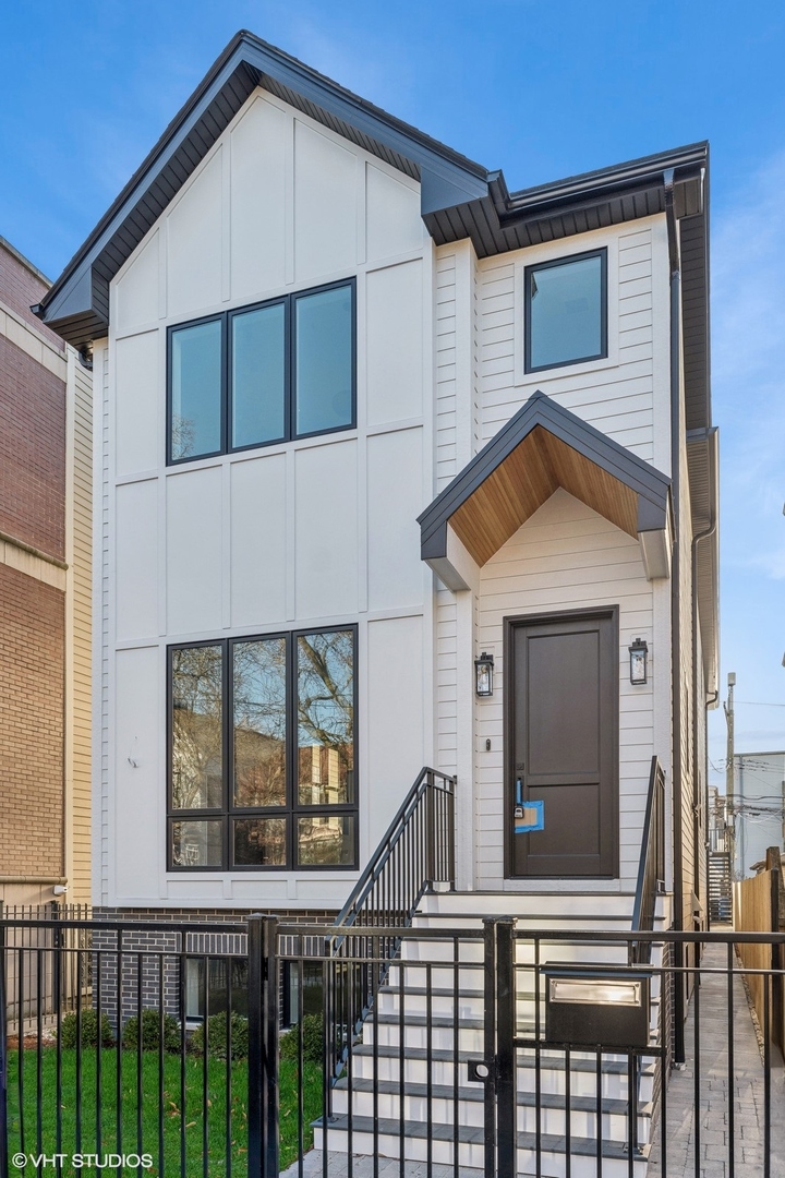 Modern white multi-story house with black-framed windows, wood-lined entry canopy, and fenced front lawn.