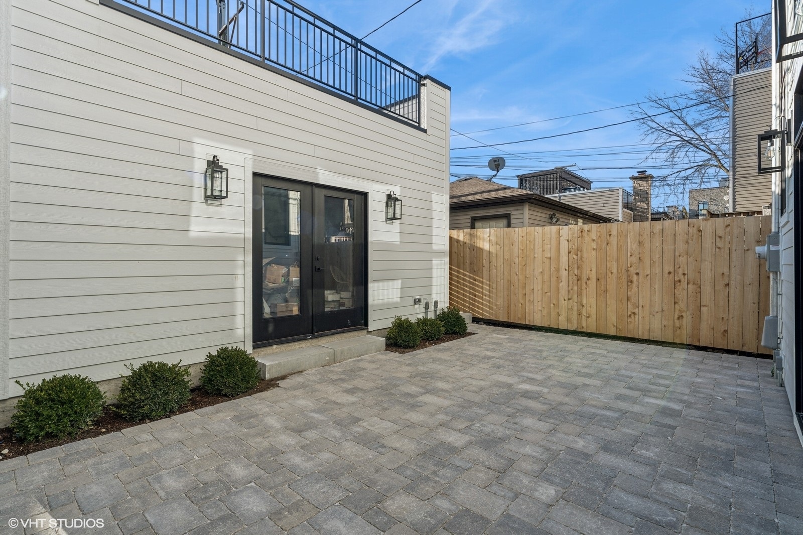 Paved backyard patio beside a white-sided home, enclosed by a tall wooden privacy fence.