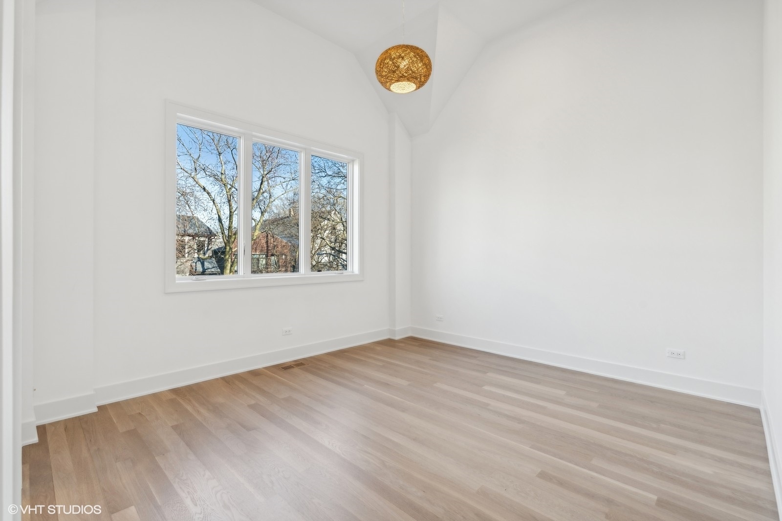 Empty room with pale wood floors, a large window, vaulted ceiling, and woven pendant light.