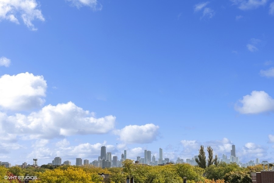 Chicago skyline rises above autumn-colored treetops beneath a blue, cloud-dotted sky.
