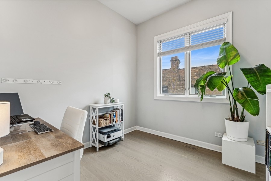 Home office with a wooden desk, white chair, bookcase, and leafy plant beside a window.